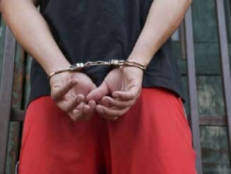 Close-up of a person in handcuffs, viewed from behind, wearing red pants and a black shirt.