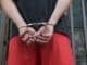 Close-up of a person in handcuffs, viewed from behind, wearing red pants and a black shirt.