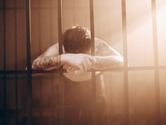 A tattooed man leans against prison bars, illuminated by dramatic sunbeams. Moody photography.