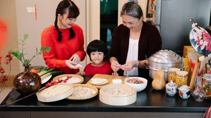 Cheerful Asian grandma with boy and female teen preparing dumplings at table with minced beef filling at home