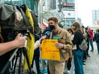 Young man holds protest sign during street demonstration against war, covered by media.
