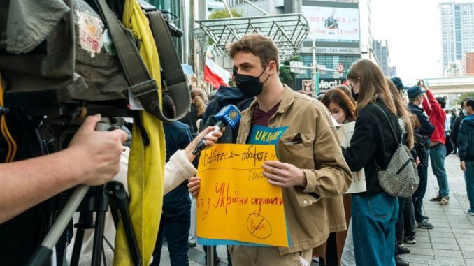 Young man holds protest sign during street demonstration against war, covered by media.
