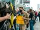 Young man holds protest sign during street demonstration against war, covered by media.