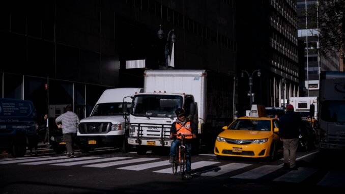 Cyclist navigates busy street in New York City traffic, surrounded by vehicles and buildings.